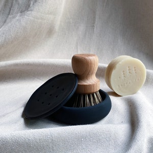 May include: A black silicone dish brush with a wooden handle and a black silicone base. A round bar of soap with the text "boxed folk" is next to the brush.