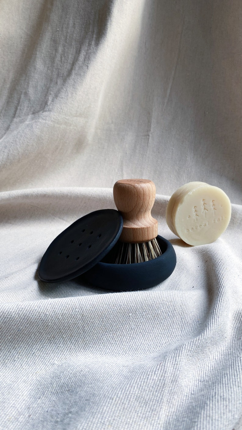 May include: A black silicone dish brush with a wooden handle and a black silicone base. A round bar of soap with the text "boxed folk" is next to the brush.