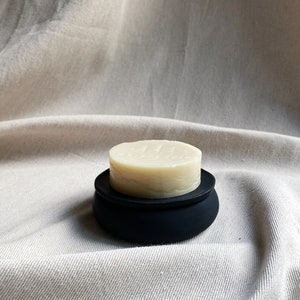 May include: A round, white soap bar in a black, round soap dish. The soap dish has a smooth, matte finish.
