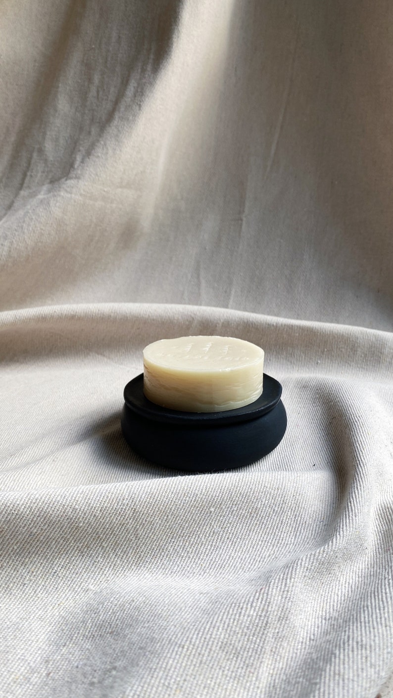 May include: A round, white soap bar in a black, round soap dish. The soap dish has a smooth, matte finish.