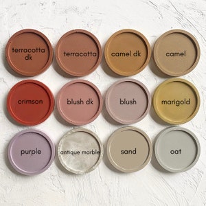 May include: Twelve round concrete coasters in various colours, including terracotta, camel, crimson, blush, marigold, purple, antique marble, sand, and oat. The coasters are arranged in three rows of four.