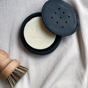 May include: A black soap dish with a white bar of soap inside. The soap has a design of trees and the text "boreal folk".