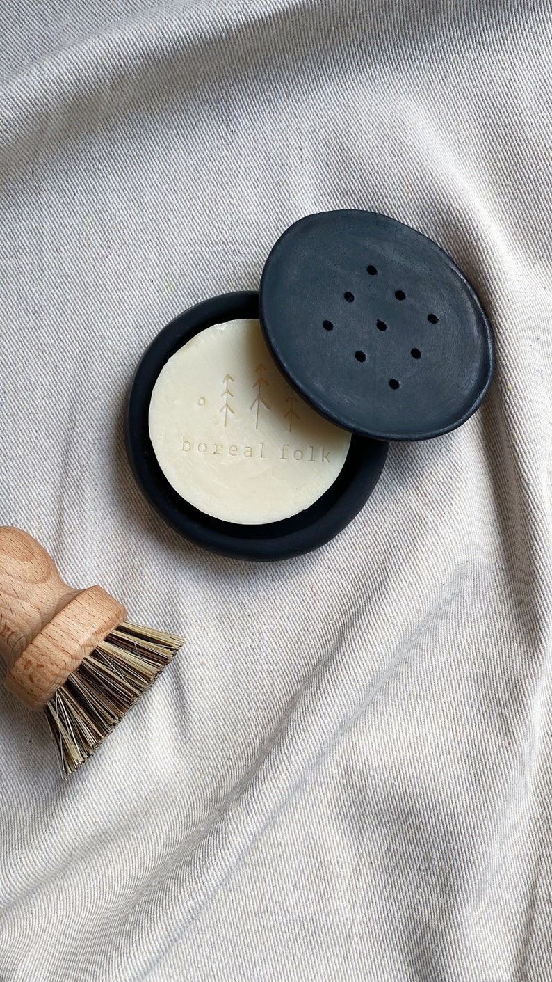 May include: A black soap dish with a white bar of soap inside. The soap has a design of trees and the text "boreal folk".