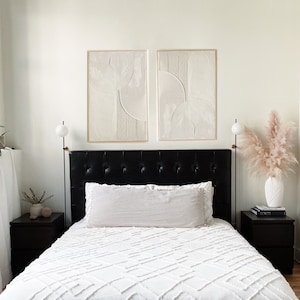 Op de afbeelding: Een wit bed met een zwart hoofdbord en een wit gewatteerd beddengoed. Twee zwarte nachtkastjes met lampen staan aan weerszijden van het bed. Twee abstracte witte en beige ingelijste kunstprints hangen aan de muur boven het hoofdbord.