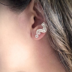 Op de afbeelding: Een zilveren oor manchet met een delicaat, draadgewikkeld ontwerp. De oor manchet buigt langs het oor, met ingewikkelde details en spiraal accenten. De sieraden worden getoond op het oor van een persoon.