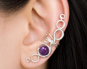 May include: A silver-toned wire ear cuff featuring a polished, round purple gemstone at the base. The design consists of intricate coiled wire loops and circular accents that extend upward along the outer edge of the ear cartilage.