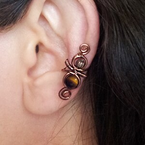 K&ouml;nnte beinhalten: Kupferfarbene Ohrklemme mit Drahtwickeldesign. Sie verf&uuml;gt &uuml;ber einen Tigeraugenstein und einen kleineren, klaren Stein. Der Schmuck wird am Ohr getragen und zeigt die filigrane Metallarbeit und die Edelsteinakzente.