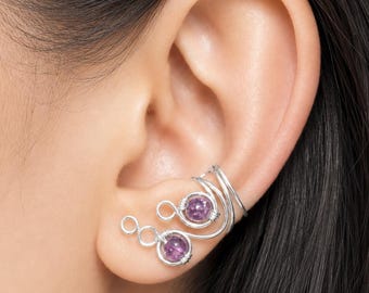 May include: Silver-toned ear cuff and earrings set. The earrings feature two purple gemstones, each set within a wire design. The ear cuff has a spiral design with a third purple gemstone. The jewelry is worn on a person's ear.