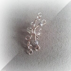 May include: A handcrafted silver wire pendant with a delicate, abstract design. The pendant features intricate wirework, including spirals and loops, and is accented with small, sparkling crystals. The overall shape is asymmetrical and artistic, suitable for jewelry.