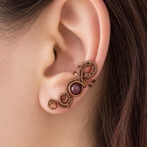 May include: A copper ear cuff with a swirling wire design and a small, round, dark red gemstone. The cuff wraps around the ear, creating a unique and artistic jewellery piece.