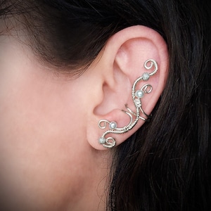 May include: A silver ear cuff with a delicate, swirling wire design. The cuff features small, round, white pearls and sparkling accents. The jewelry is worn on the ear, showcasing an elegant, nature-inspired aesthetic.