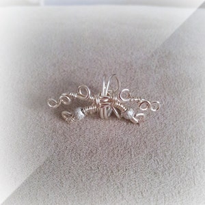May include: A handcrafted silver-toned wire wrap jewelry piece. The design features intricate wire weaving with decorative silver beads. The piece has a delicate, artistic appearance, suitable for a unique accessory.