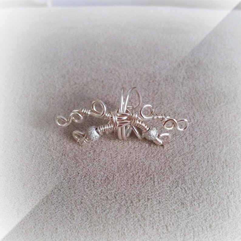 May include: A handcrafted silver-toned wire wrap jewelry piece. The design features intricate wire weaving with decorative silver beads. The piece has a delicate, artistic appearance, suitable for a unique accessory.