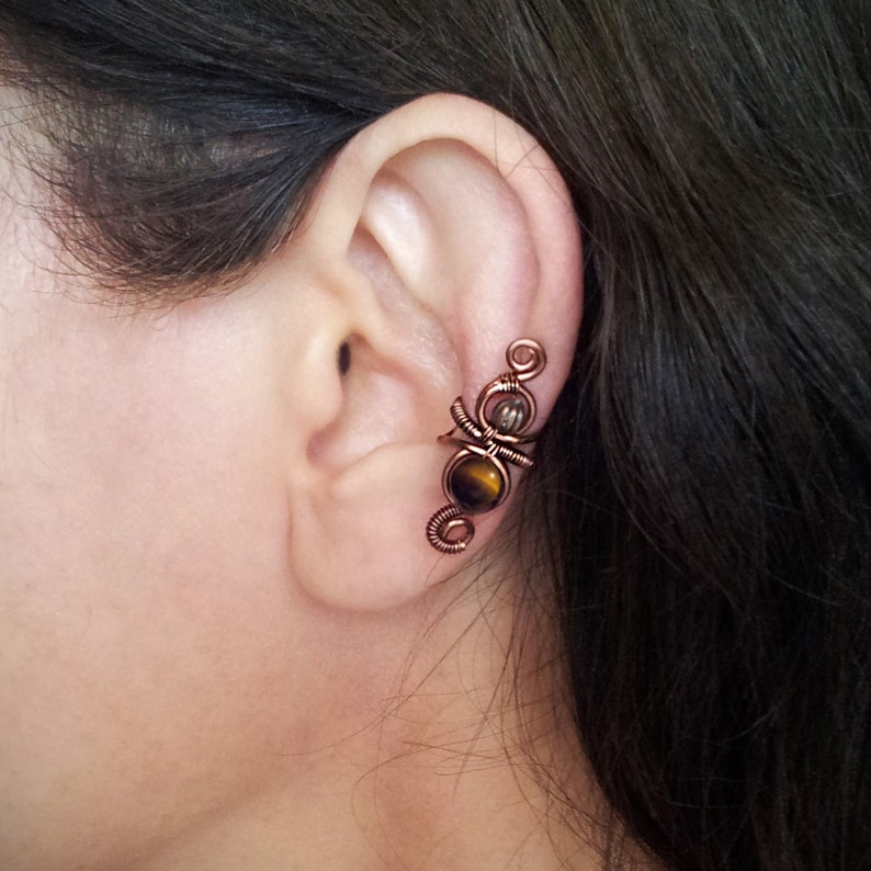 K&ouml;nnte beinhalten: Kupferfarbene Ohrklemme mit Drahtwickeldesign. Sie verf&uuml;gt &uuml;ber einen Tigeraugenstein und einen kleineren, klaren Stein. Der Schmuck wird am Ohr getragen und zeigt die filigrane Metallarbeit und die Edelsteinakzente.