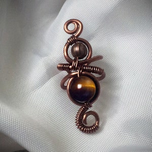 K&ouml;nnte beinhalten: Ein handgefertigter Kupferanh&auml;nger mit einem wirbelnden Drahtdesign. Der Anh&auml;nger hat einen Tigeraugen-Edelstein und eine kleinere braune Perle, die alle kunstvoll in Kupferdraht gewickelt sind. Der Anh&auml;nger ist etwa 5 cm lang.