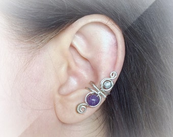 May include: A silver-toned ear cuff featuring a wire-wrapped design. The cuff has a round, purple gemstone and a small, clear stone. The design curves along the ear, with decorative swirls and wrapped wire details.