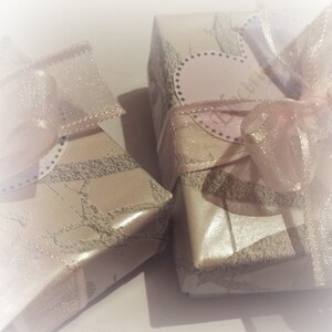 May include: Two small, rectangular gift boxes wrapped in patterned paper with a light beige background and a gray design. Each box is tied with a sheer, light pink ribbon and bow. One box has a pink heart-shaped tag.
