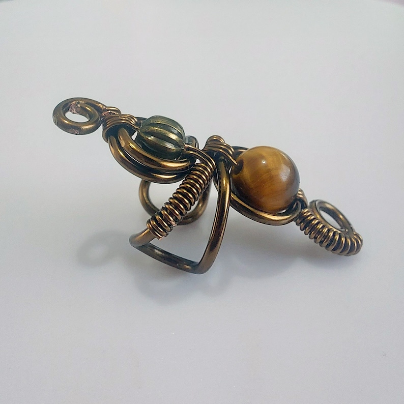 K&ouml;nnte beinhalten: Handgefertigter Ring aus bronzefarbenem Draht mit einem Tigeraugen-Perle und einer gr&uuml;nen, geriffelten Perle. Der Ring hat ein einzigartiges, k&uuml;nstlerisches Design mit gewendelten Drahtakzenten und einer dekorativen Schlaufe.