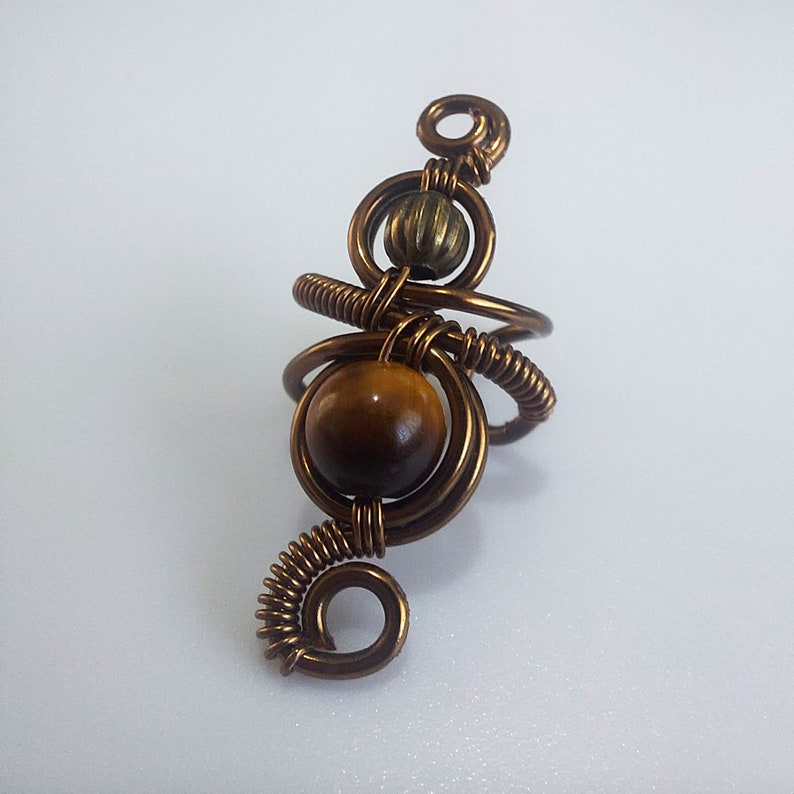 K&ouml;nnte beinhalten: Handgefertigter Kupferdrahtring mit einem gro&szlig;en, runden, braunen Tigerauge-Stein. Der Ring hat ein einzigartiges, k&uuml;nstlerisches Design mit spiralf&ouml;rmigem Draht und einer kleinen, strukturierten Perle. Der Ring ist vor einem wei&szlig;en Hintergrund platziert.