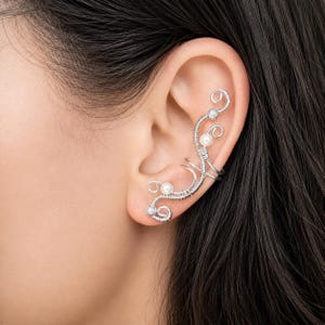 Könnte beinhalten: Ein silberner Ohrklemme mit einem filigranen Drahtdesign. Es verfügt über kleine, runde Perlen und klare Kristalle. Das Design schmiegt sich an das Ohr an und schafft ein elegantes Accessoire.