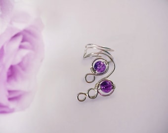 May include: A silver-toned ear cuff featuring two purple gemstone beads. The wire-wrapped design curves around the ear, with a decorative loop at the bottom. The cuff is set against a soft, blurred background.