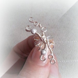 May include: A silver-colored wire ring with a unique, abstract design. The ring features intricate wire wrapping, small beaded accents, and decorative swirls. The ring is held in a hand, showcasing its delicate craftsmanship and artistic flair.