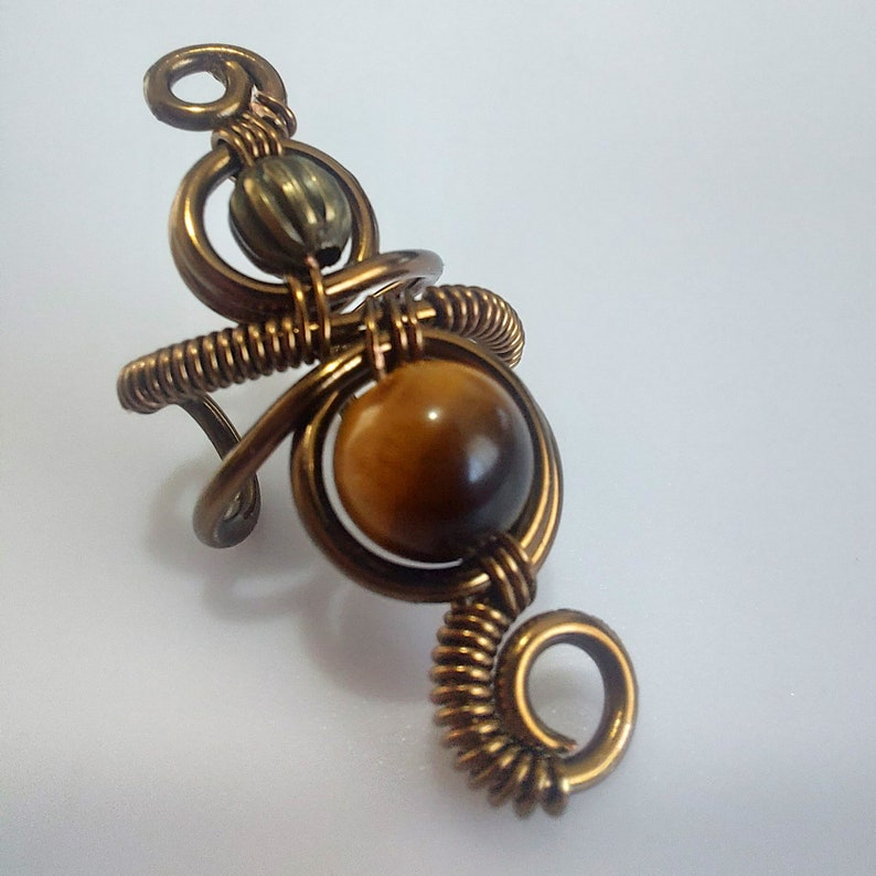 K&ouml;nnte beinhalten: Handgefertigter Kupferdrahtring mit einem gro&szlig;en, runden Tigeraugenstein und einer kleineren, strukturierten Perle. Der Ring hat ein einzigartiges, k&uuml;nstlerisches Design mit aufwendiger Drahtwicklung und einem Spiraldetail.