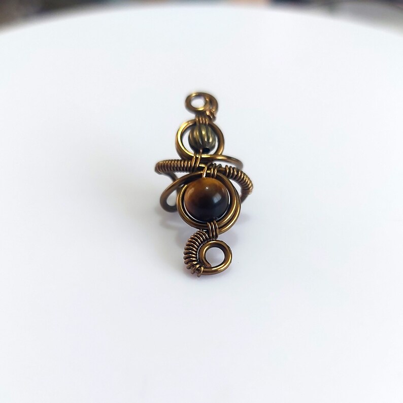 K&ouml;nnte beinhalten: Ein handgefertigter, kupferfarbener Drahtwickelring mit einem runden, polierten Tigeraugen-Edelstein. Der Ring hat ein einzigartiges, k&uuml;nstlerisches Design mit aufwendigen Drahtdetails und einem Spiralakzent. Der Edelstein hat einen Durchmesser von ca. 1,3 cm.