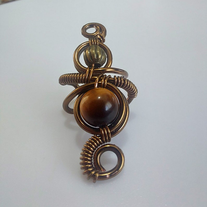 K&ouml;nnte beinhalten: Handgefertigter Kupferdrahtring mit einem gro&szlig;en, runden, braunen Tigeraugenstein. Der Ring hat ein einzigartiges, k&uuml;nstlerisches Design mit gewickelten und geschlungenen Drahtelementen und einer kleinen, strukturierten Perle.