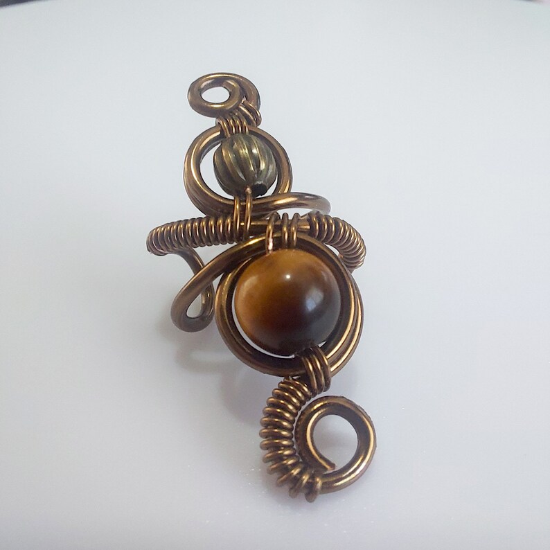 K&ouml;nnte beinhalten: Handgefertigter kupferfarbener Drahtring mit einem gro&szlig;en, runden Tigeraugenstein. Der Ring hat ein aufwendiges, k&uuml;nstlerisches Design mit einer kleineren, strukturierten Perle und gewendelten Drahtakzenten. Ein einzigartiges Schmuckst&uuml;ck.