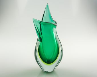 May include: A green glass vase with a unique, abstract design. The vase has a wide mouth and a narrow base, and it is made of thick, clear glass. The vase is perfect for displaying flowers or other decorative items.