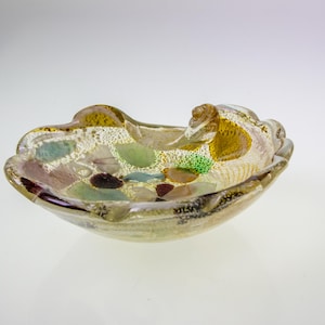Op de afbeelding: Een kleine, witte glazen kom met een gegolfde rand. De kom is versierd met verschillende gekleurde glazen stukjes, waaronder groen, bruin en goud.