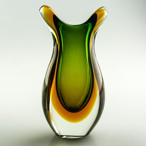 Könnte beinhalten: Eine klare Glasvase mit einer einzigartigen Form und einer Mischung aus Farben. Die Vase hat einen leuchtend grünen Innenraum, Übergänge zu orangefarbenen und gelben Akzenten und eine klare Außenseite. Der obere Teil hat zwei ausgestellte Öffnungen.