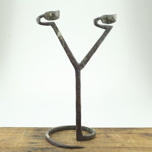 May include: A handcrafted, wrought iron candle holder with a Y-shaped design. The candle holder has two small, cup-shaped holders at the top and a coiled base. The metal has a rustic, aged appearance, with visible texture and color variations.