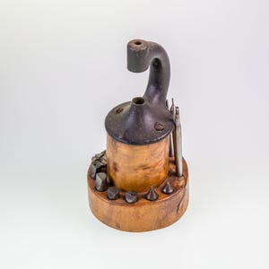 May include: A vintage, wooden tool holder with a cylindrical body and a curved, black metal top. The base features various metal bits and tools. The tool holder is a light brown color, with a dark brown top and accents.
