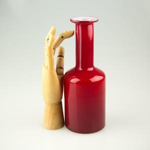 Op de afbeelding: Een hoge, rode glazen vaas met een afgeronde basis en een smalle hals. De vaas heeft een glanzende afwerking en een lichtere rode rand. Een houten, gelede hand staat naast de vaas.