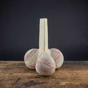 Op de afbeelding: Een keramische vaas met een lange, slanke hals en drie ronde, bolvormige bases. De vaas is versierd met een geometrisch patroon van roze en witte lijnen.