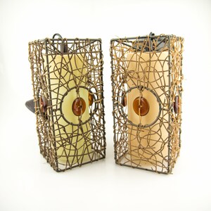 Op de afbeelding: Twee decoratieve kandelaars. Elke houder heeft een rechthoekige, geweven metalen kooi in een bruine tint, met een crèmekleurige kaars erin. Amberkleurige knopen en cirkelvormige accenten sieren de voorkant. De houders zijn ontworpen voor woondecoratie.