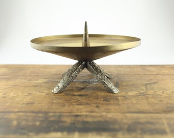 May include: A vintage brass candle holder with a conical base and three textured legs. The candle holder has a wide, shallow dish and a central spike for securing a candle. The metal has a warm, aged patina, suggesting its antique nature.
