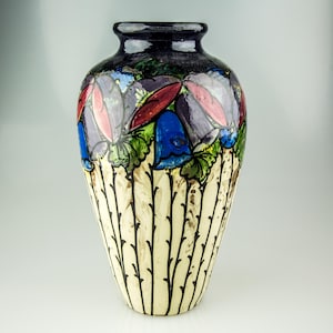 Puede incluir: Un jarrón decorativo con base crema y borde azul oscuro. El jarrón presenta un diseño floral con flores rojas, azules y moradas. Líneas verticales negras adornan la base. Una pieza única para la decoración del hogar.