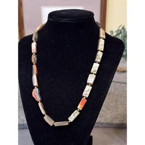 May include: A necklace with rectangular beads in shades of beige, orange, gray, and iridescent white. The beads are strung together with small gold-colored spacers. The necklace is displayed on a black mannequin.