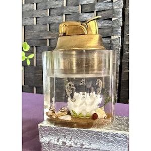 May include: A vintage lighter with a gold-toned top and a clear, cylindrical base. The base features a miniature underwater scene with seahorses, coral, shells, and sand. The lighter is likely from the mid-20th century.
