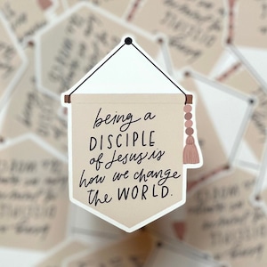 May include: A beige and brown banner with a pink tassel hanging from a string. The banner reads "being a DISCIPLE of Jesus is how we change the WORLD."