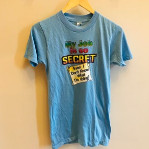 May include: A light blue t-shirt with a rainbow graphic that says "My Job is so SECRET" and a yellow note that says "Even I Don't Know What I'm Doing!"