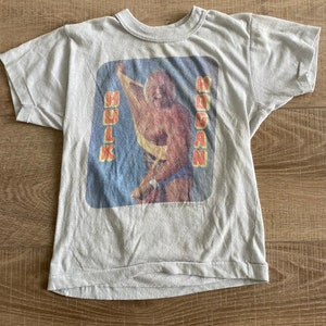 May include: A gray t-shirt with a vintage image of Hulk Hogan. The image shows Hogan in a wrestling pose, with the words "Hulk Hogan" printed on the shirt.