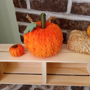 May include: A plush orange pumpkin with a green stem and brown twine accents. The pumpkin is sitting on a wooden shelf with a smaller orange pumpkin in the background.