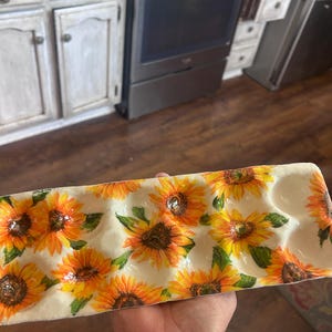 May include: A rectangular ceramic egg holder decorated with vibrant sunflowers. The holder has a cream-coloured base and features multiple depressions for holding eggs. The sunflowers are painted with yellow, orange, and brown hues, with green leaves.