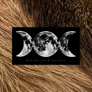 May include: A black and white image of a triple moon symbol, with the full moon in the center and crescent moons on either side. The image is on a black background and has the text "@julia.rose.waters" at the bottom.