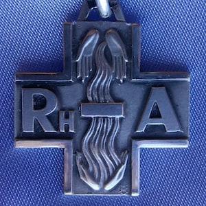 May include: A silver cross pendant featuring the letters "RHA" and a flame motif. The pendant is suspended from a loop. The backdrop is a blue fabric, providing a contrasting background.