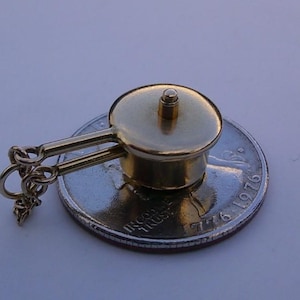 Peut inclure: Un petit charme en forme de casserole dorée avec un couvercle et deux poignées. Le charme est attaché à une chaîne et repose sur une pièce de monnaie. Le charme est probablement un bijou.